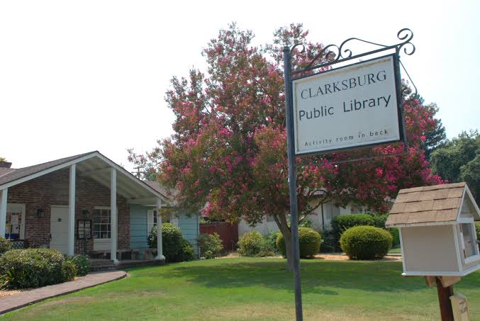 Exterior of Clarksburg Public Library, a single-story brick building with a covered entrance, landscaped lawn, and flowering trees. A sign identifies the library and notes that the activity room is in the back.