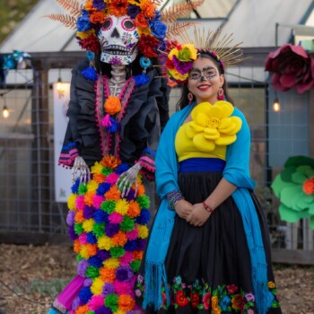 Two people in colorful Día de Muertos attire and face paint, posing together at a community celebration.