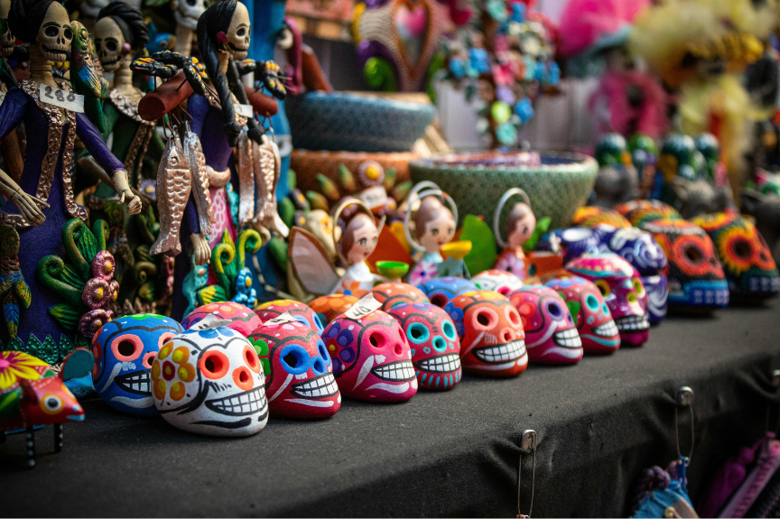 Colorful Día de los Muertos figurines displayed at a market, including painted skulls with floral patterns and grinning faces, skeleton figures, and small dolls arranged on a table.