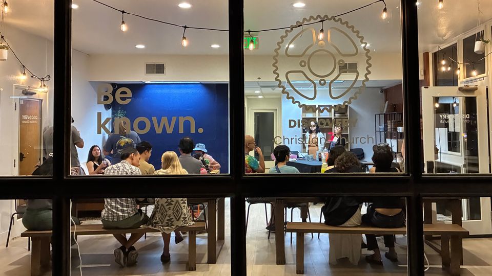 People gathered around tables inside Discovery Hub, viewed through large front windows displaying the words “Be known” and “Discover Christian Church.” Warm string lights hang from the ceiling.