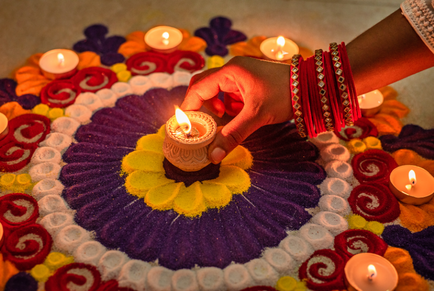 A hand wearing red and gold bangles places a lit clay oil lamp in the center of a colorful floral rangoli surrounded by glowing candles, representing the Diwali festival of lights.