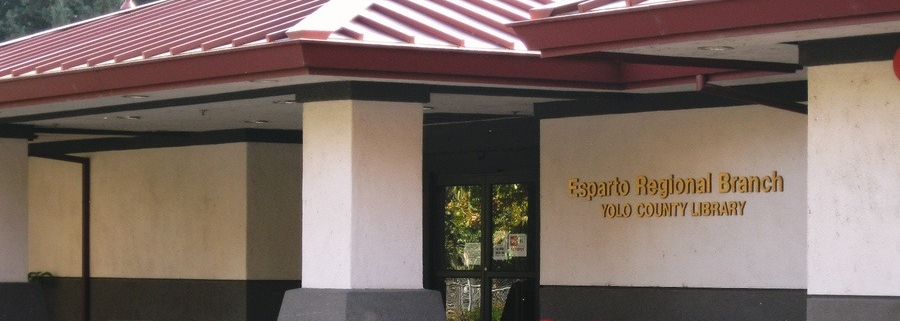 Exterior of the Esparto Regional Branch of the Yolo County Library, featuring a red metal roof, light-colored walls, dark lower trim, and a recessed glass entrance.