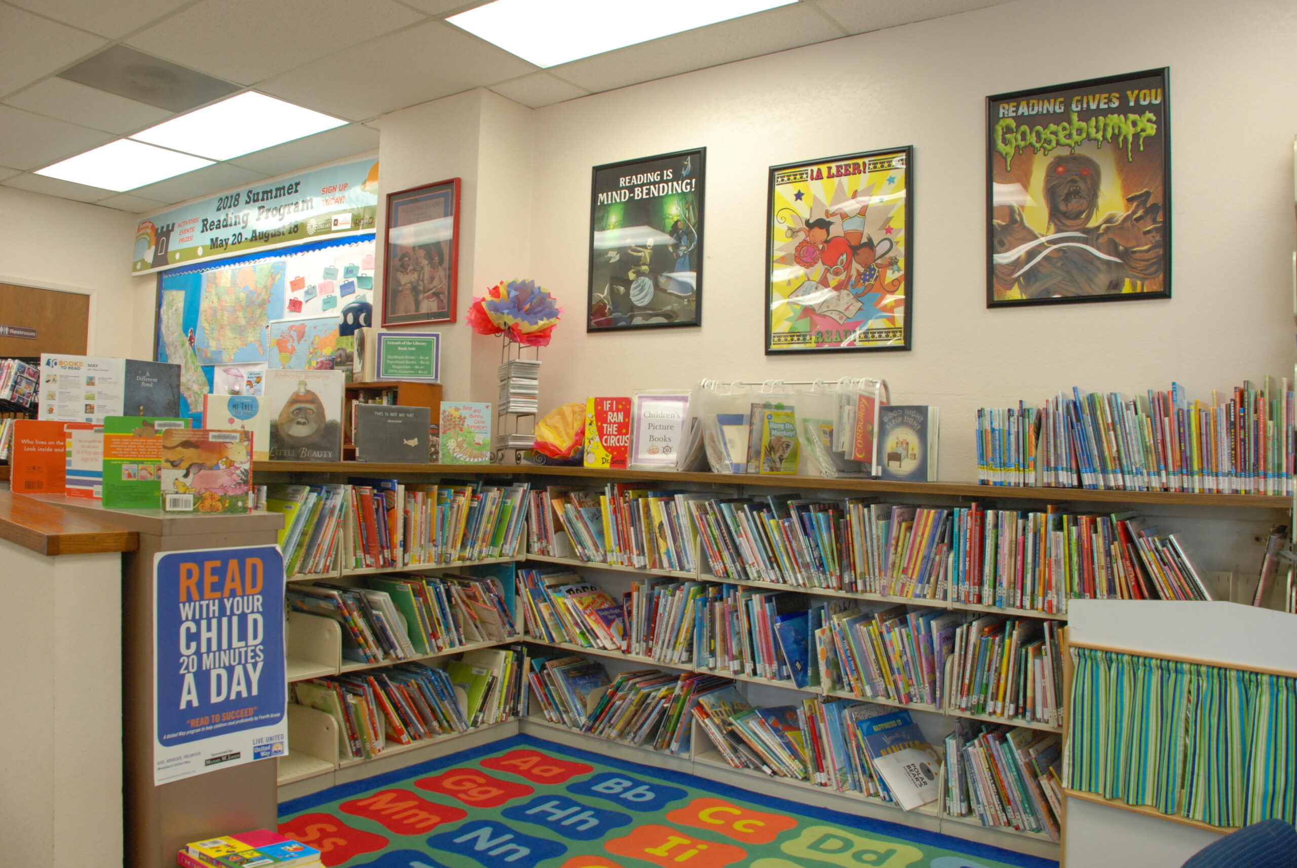Children’s section of the Knights Landing Branch Library featuring shelves filled with colorful picture books, a bright alphabet rug, framed reading-themed posters, and a sign encouraging parents to read with their children for 20 minutes a day.