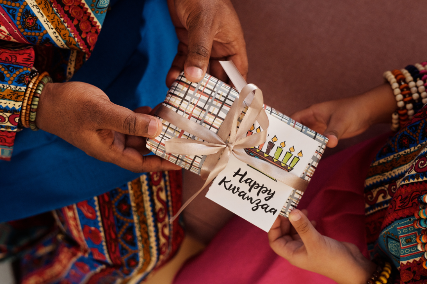 Two people exchange a wrapped gift decorated with a ribbon and a card reading “Happy Kwanzaa,” featuring a drawing of seven candles representing the Kwanzaa kinara.