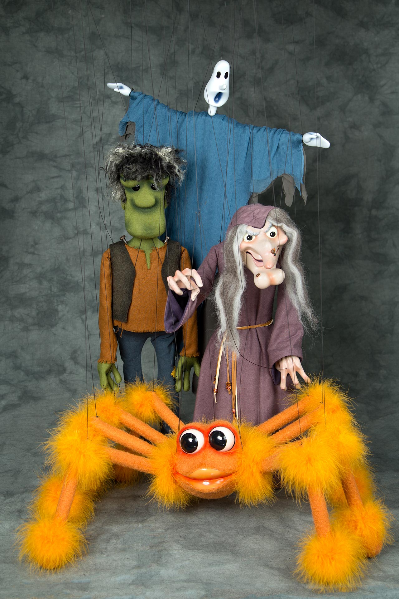Four Marinette puppets dressed as Halloween characters: a ghost in a blue draped costume, Frankenstein’s monster in an orange shirt and dark vest, a witch with long gray hair and a purple robe, and a large orange, furry spider. Puppet strings are visible against a gray backdrop.