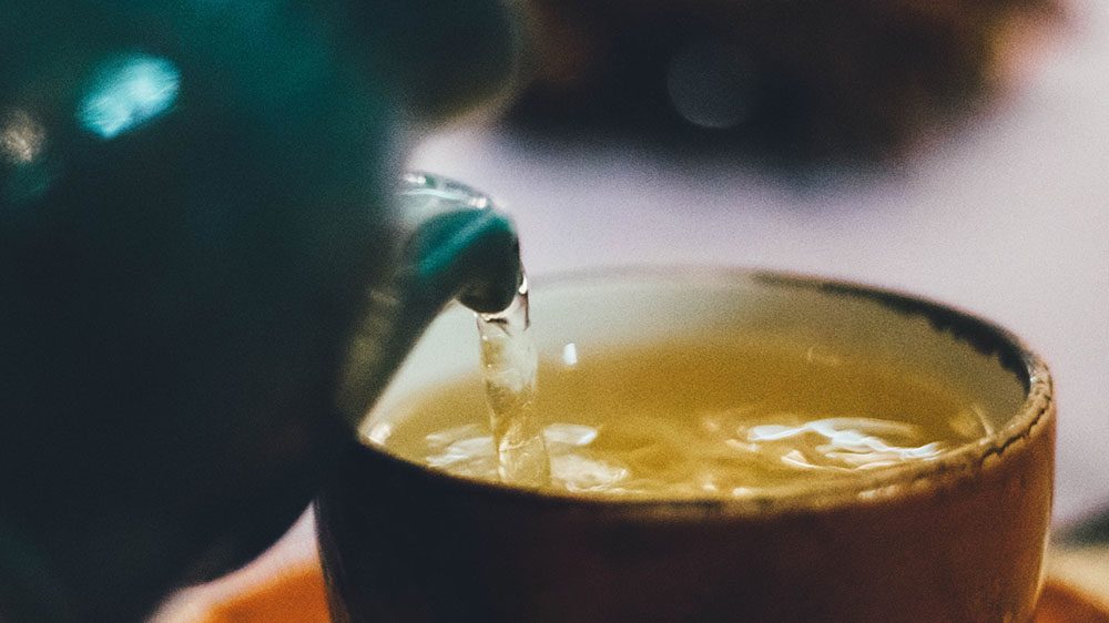 A teal kettle pours hot water into a brown tea bowl