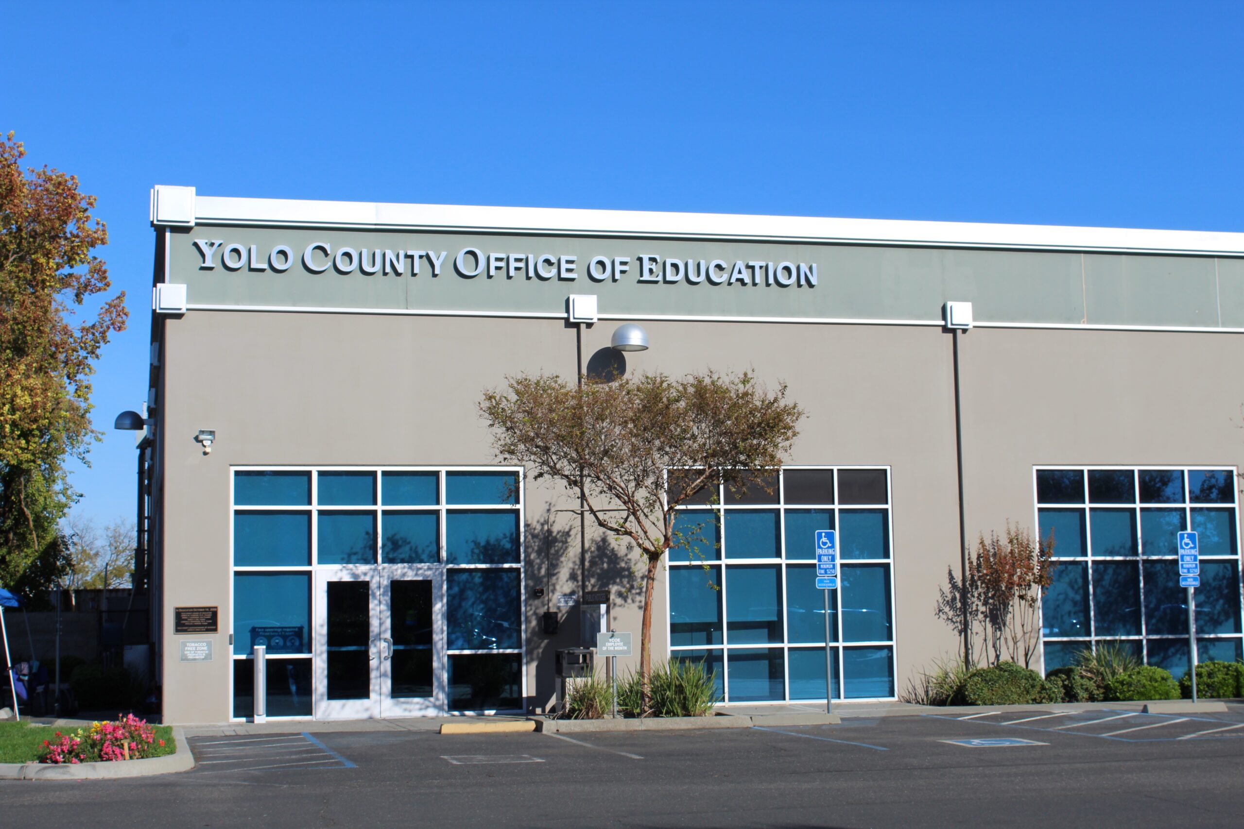 Exterior view of the Yolo County Office of Education building, featuring the organization’s name above large glass windows and entrance doors, landscaped shrubs, and accessible parking spaces in front.